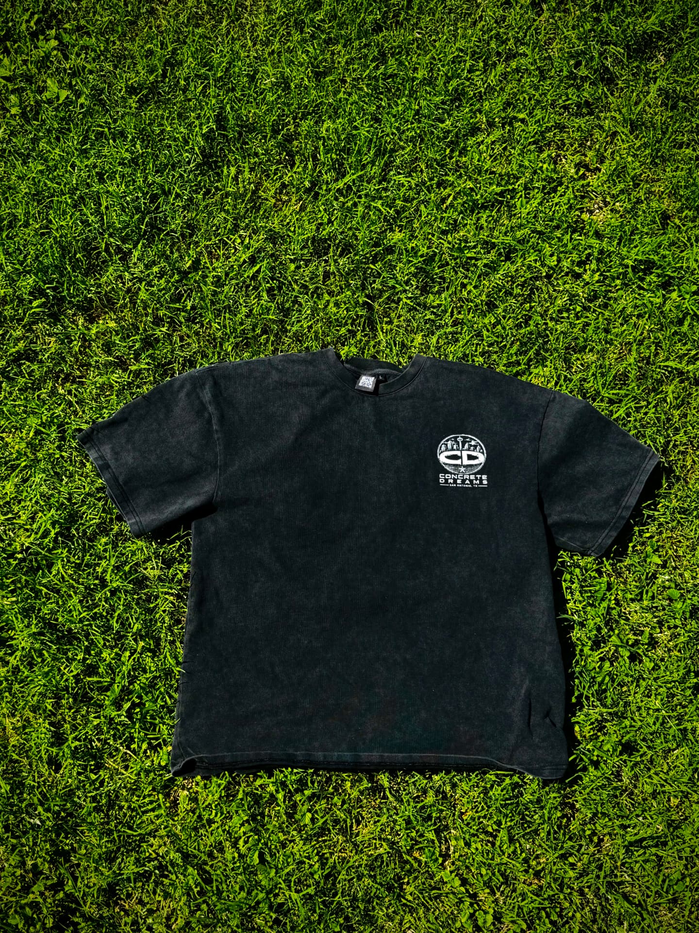 Washed black boxy short sleeve tee with a small white circular CD city logo on the left chest, laid flat on green grass